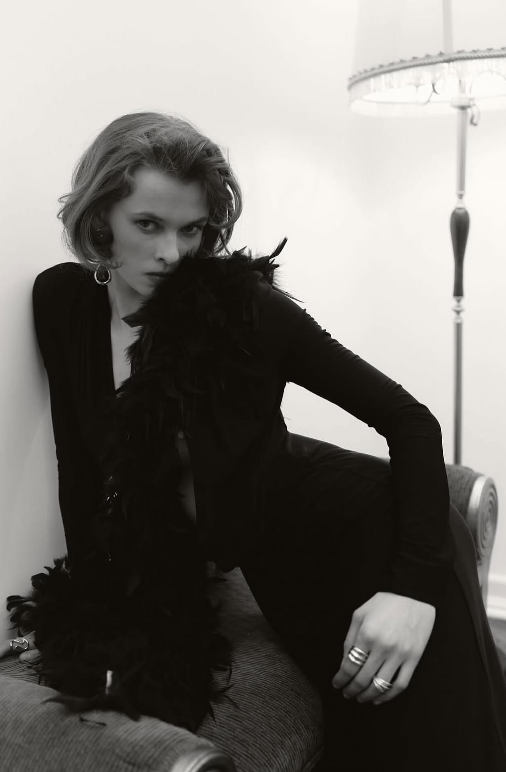 Black-and-white: Mariia in a black dress and feather boa, leaning on an armchair beside a floor lamp